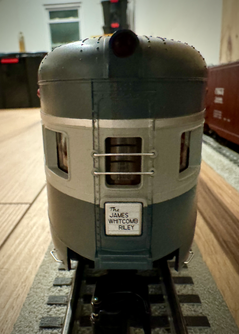 The image shows the rear end of a model train car, specifically a passenger car. The car is painted in a two-tone color scheme, with the top half being a darker color and the bottom half a lighter color. The windows have silver framing, and the back of the train car features a small sign that reads 'The JAMES WHITCOMB RILEY.' The model train is situated on a track, and the image captures the details of the car, including its rounded rear end and the texture of the paint.