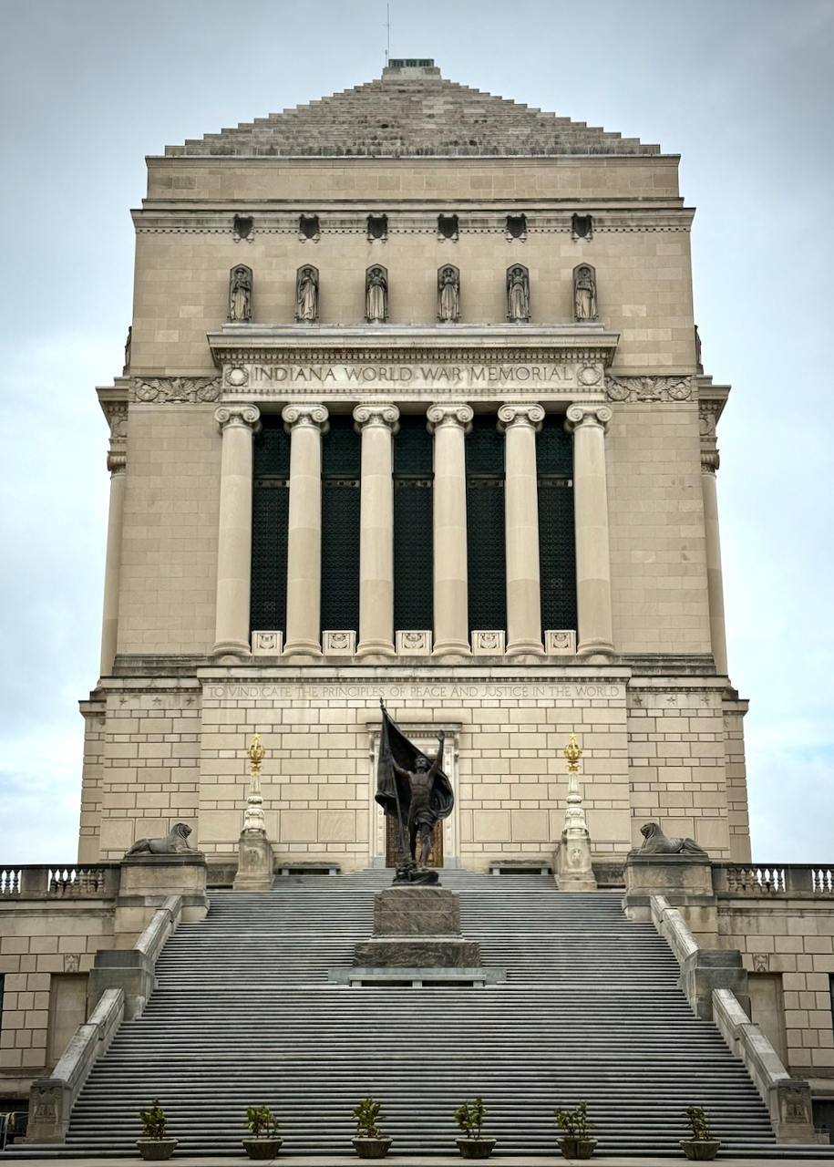 The image shows the Indiana World War Memorial, a large, rectangular monument with neoclassical architectural elements. Its inscription reads 'Indiana World War Memorial' above a row of statues and columns. In front of the monument, a staircase leads up to a bronze sculpture titled Pro Patria, which depicts a soldier holding a flag, flanked by two stone sphinxes.