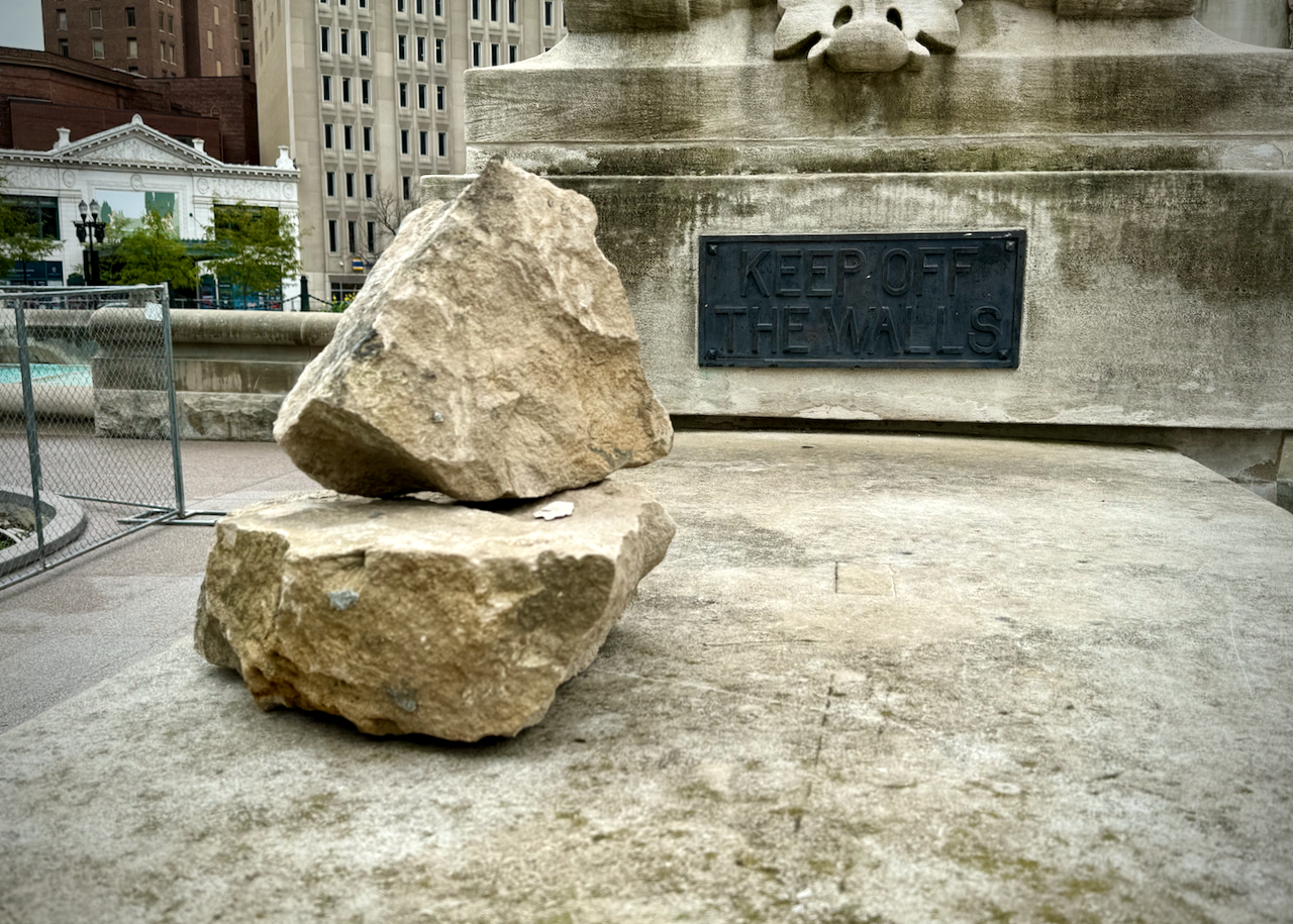 The image shows a close-up of a stone wall of the Soldiers' and Sailors' Monument in Indianpaolis. On the ledge are two large, stacked rocks. A metal plaque is affixed to the wall behind the rocks which reads, 'KEEP OFF THE WALLS,' as a warning. On the left you can see the Hilbert Circle Theatre and the AES Indiana headquarters.
