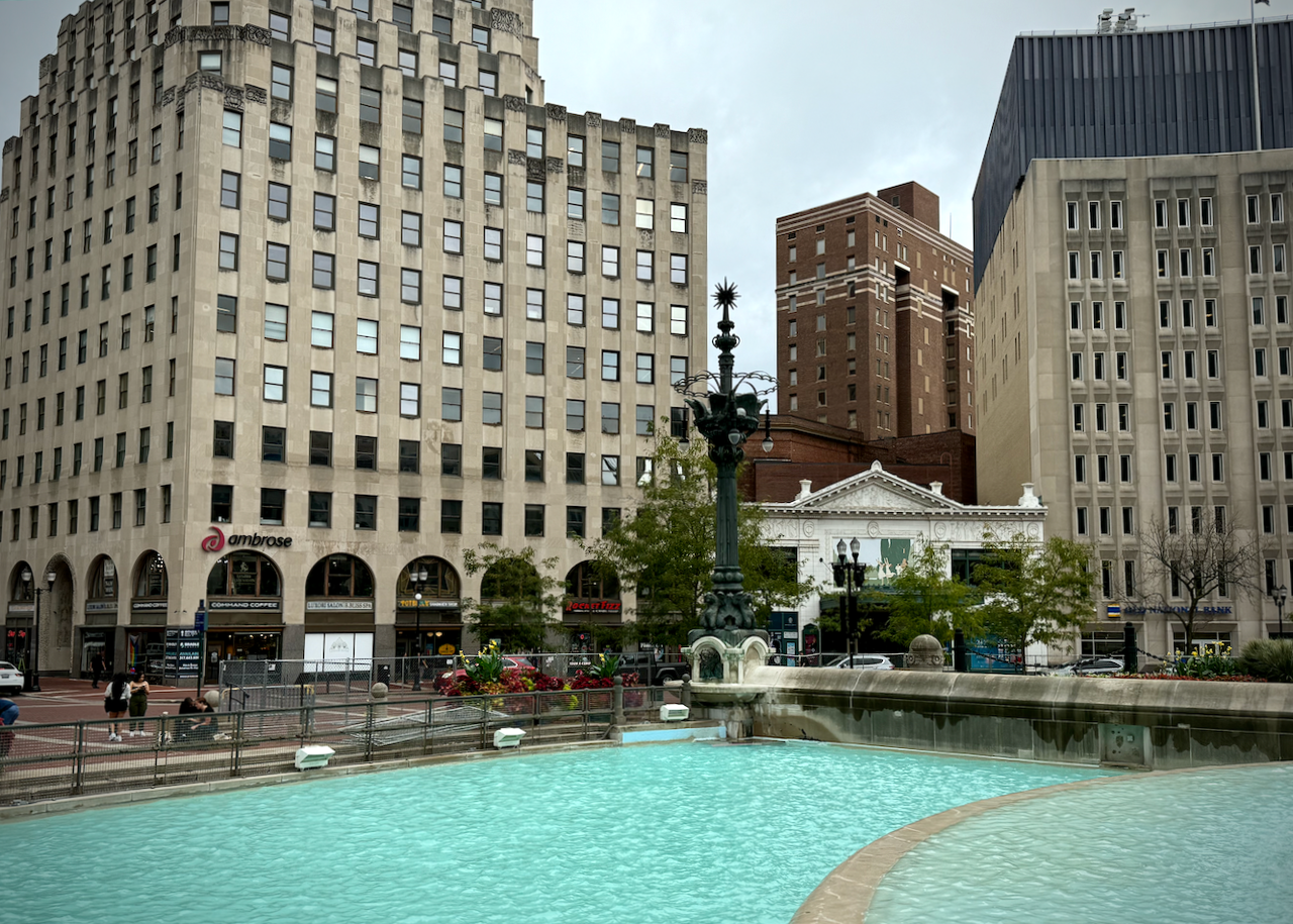 View of the southeast quadrant of Monument Circle, with a prominent water fountain pool in the foreground, part of the Soldiers' and Sailors' Monument. Across the street from the pools are several tall buildings, including an Art Deco high-rise on the left with a sign that says 'ambrose' on it. To the right is the Hilbert Circle Theatre, and then the headquarter of AES Indiana. The tall brick building in the background is Symphony Center.