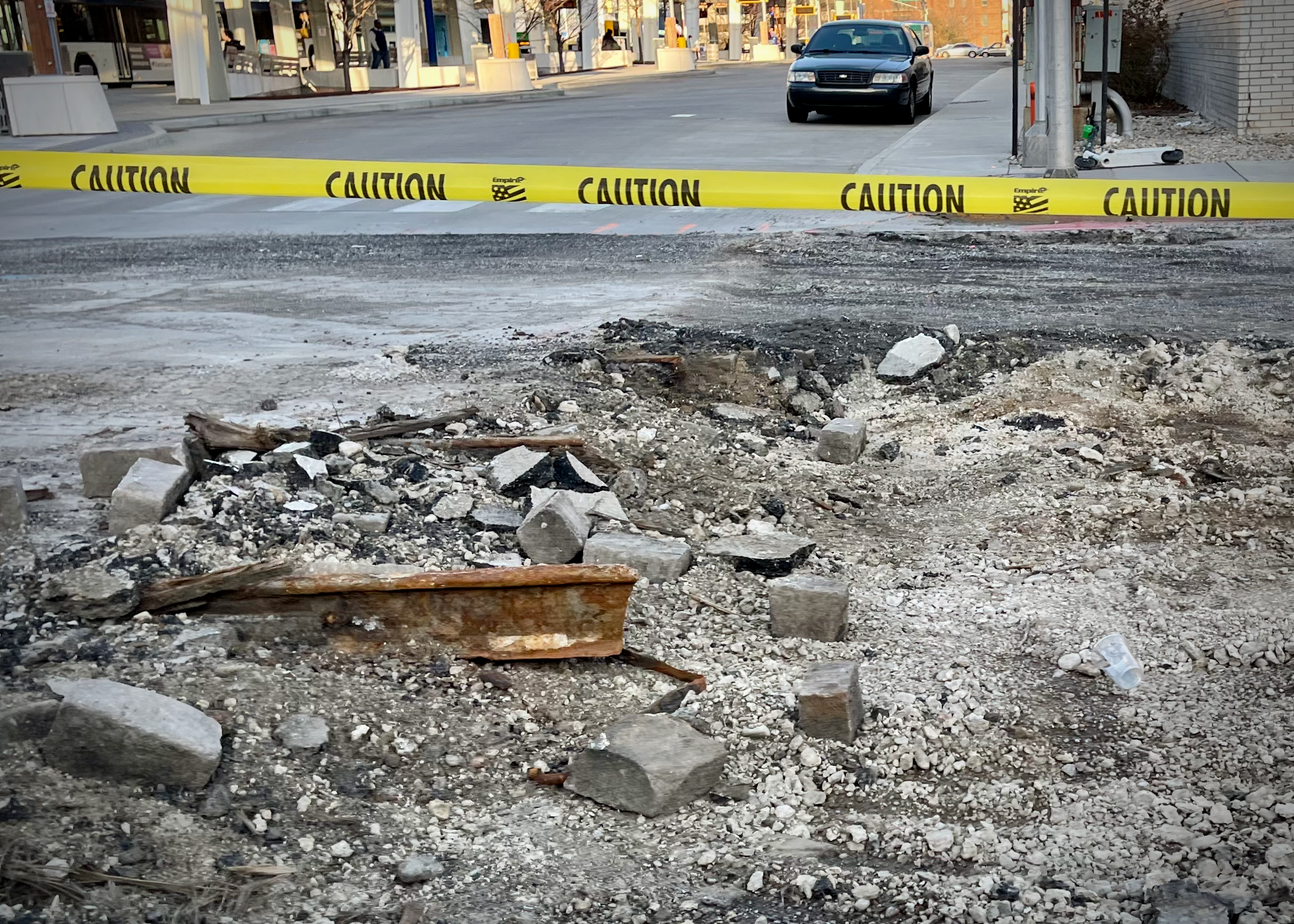 A torn-up street corner roped off with yellow caution tape reveals chunks of concrete, stone blocks, and a rusted rail segment left behind from a removed streetcar line.