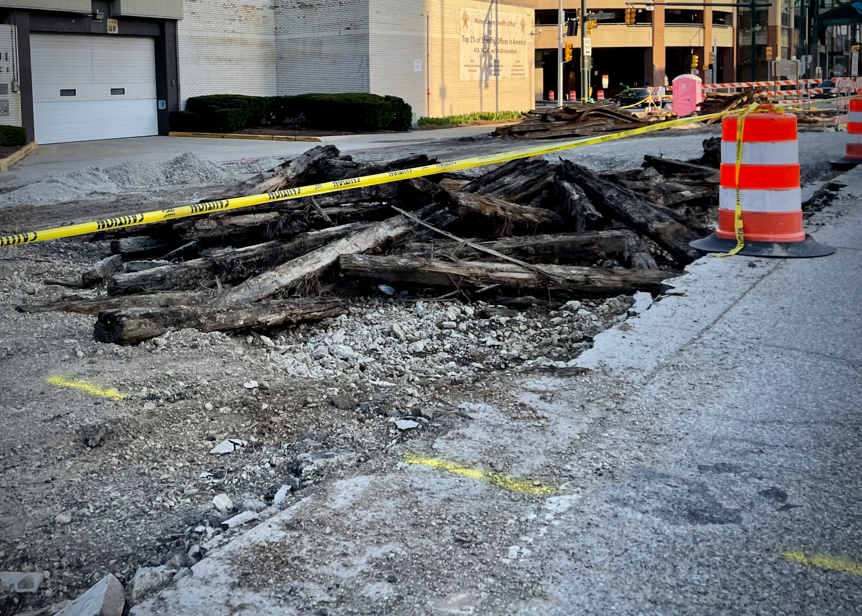 A construction zone with caution tape and orange barrels surrounds a pile of rotted wooden ties and rubble, marking the removal of an old streetcar line from the torn-up roadbed.