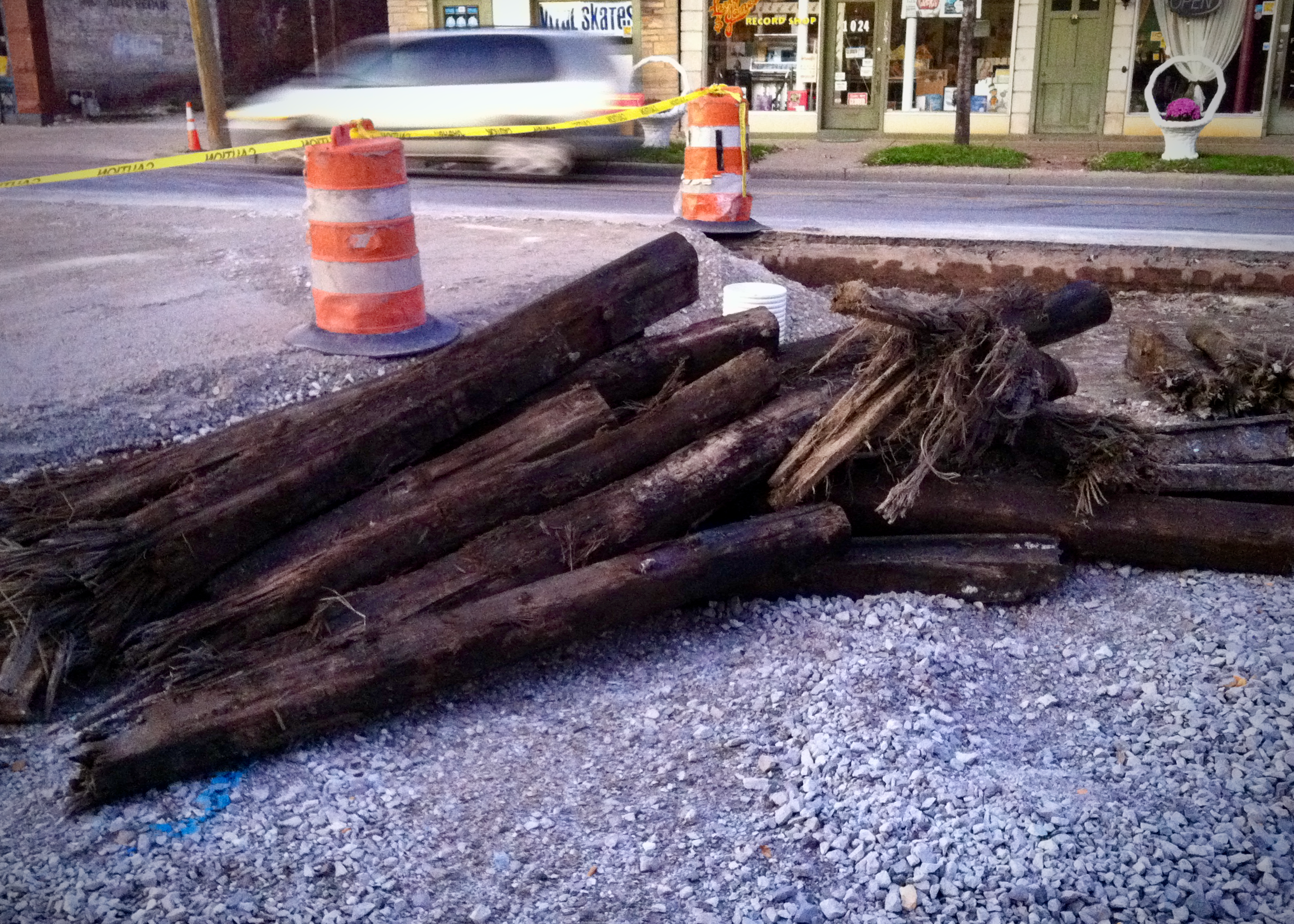 A close view of stacked, decaying wooden ties beside a street under construction, showing remnants of a dismantled streetcar line amid gravel and safety cones.