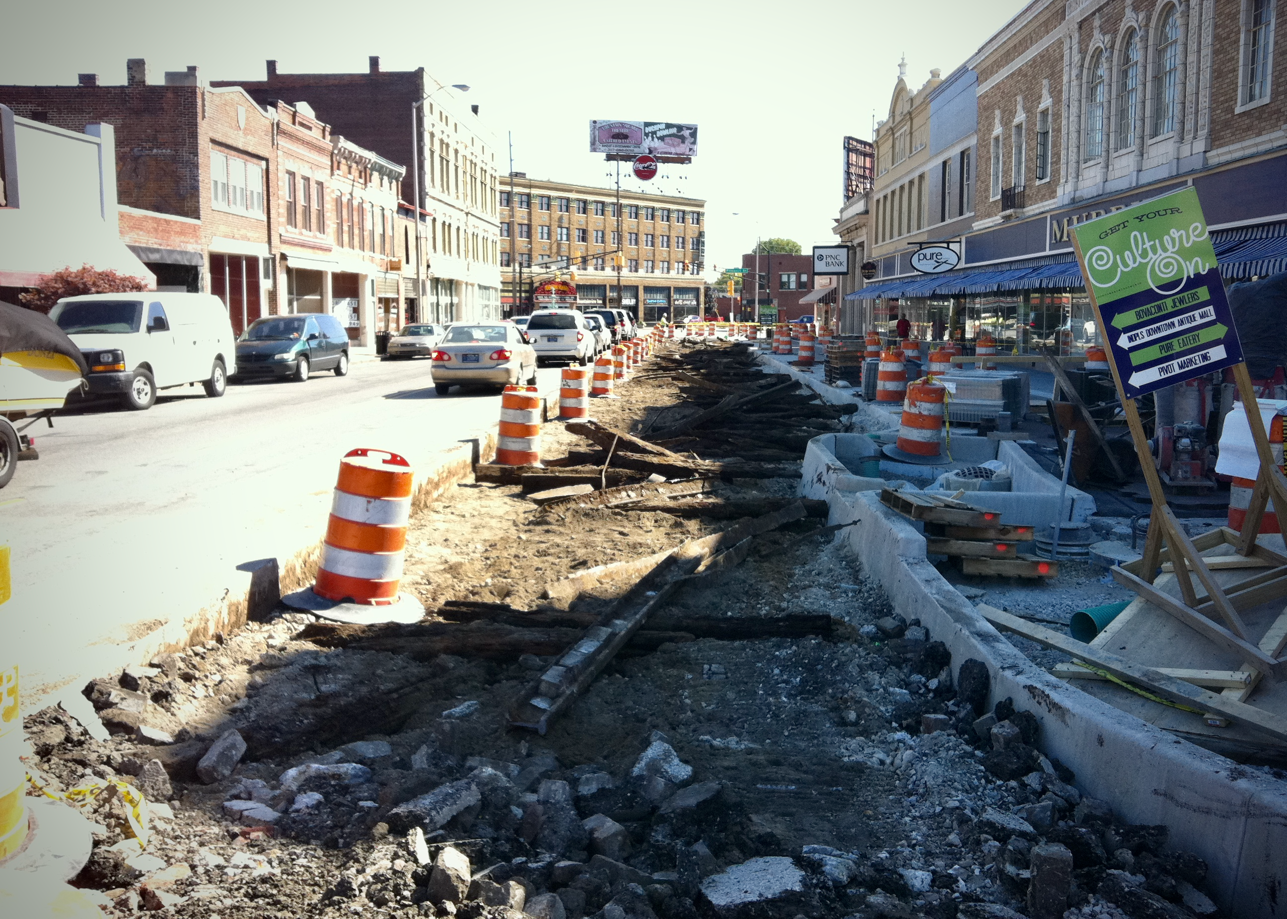 A wide view of a commercial street under construction, with dozens of orange barrels lining the sidewalk and a long path of unearthed, discarded streetcar ties extending into the distance.